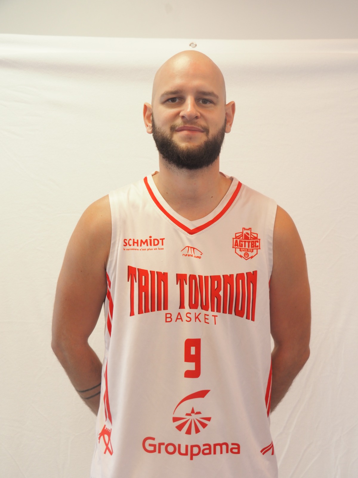 Homme chauve avec barbe portant un maillot de basket blanc et rouge numéro 9 de Trin Tournon.