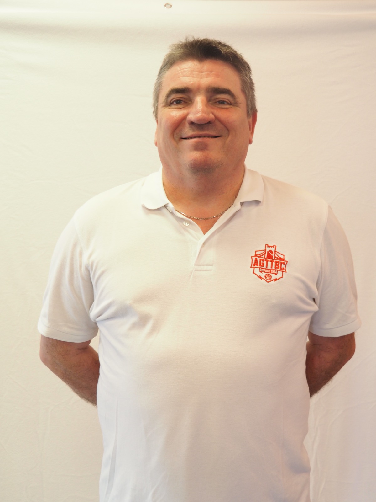 Homme souriant portant un polo blanc avec un logo rouge AGTIBC, fond blanc neutre.