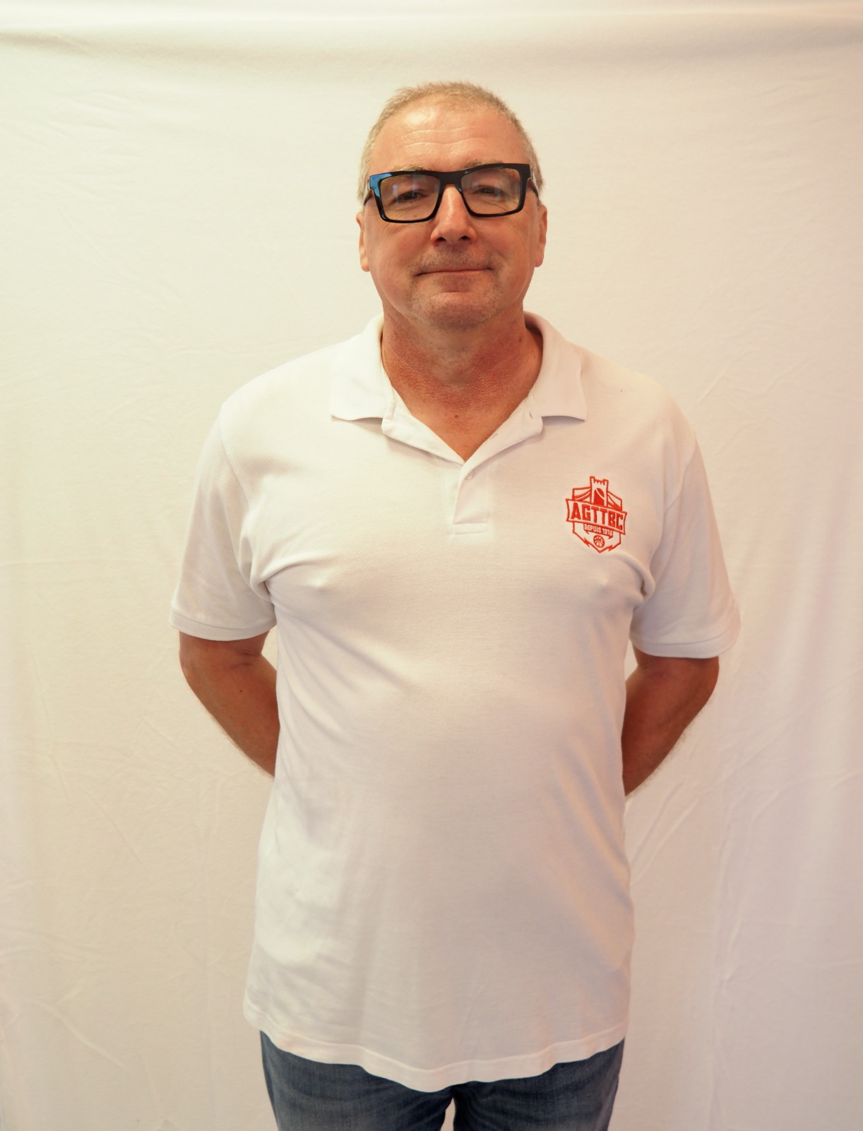 Homme portant un polo blanc avec un logo rouge AGITBC, debout devant un fond blanc.