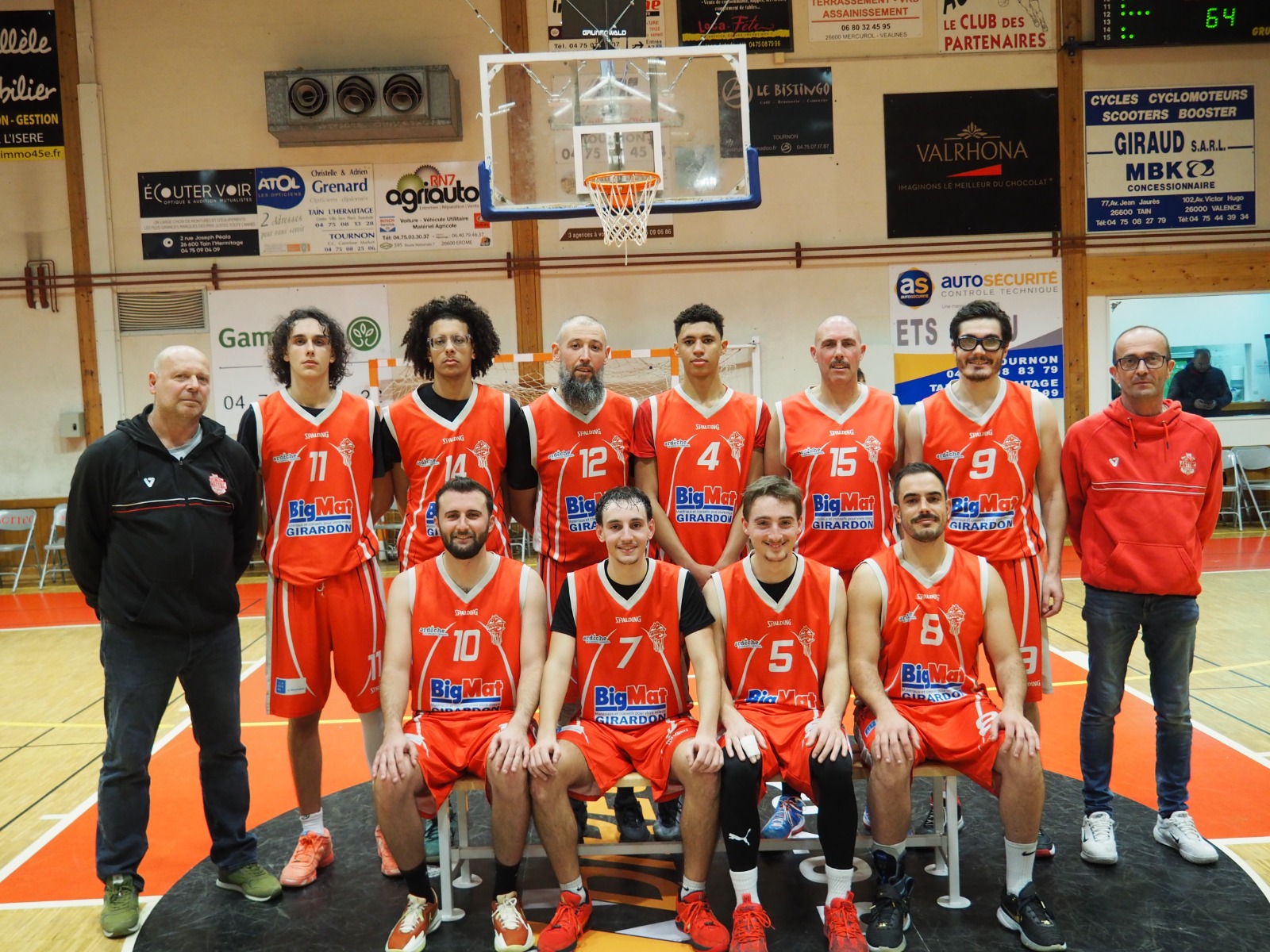 Équipe masculine de basketball en maillots orange posant sur le terrain avec deux entraîneurs.