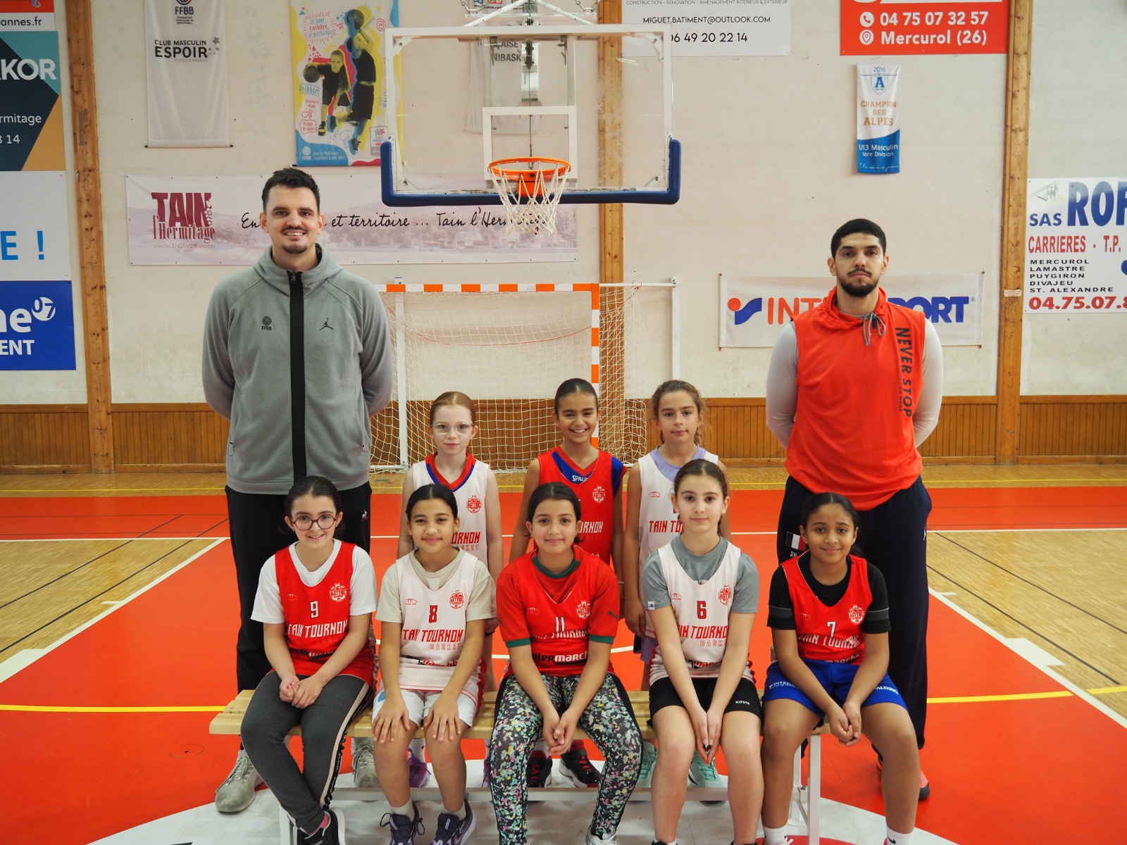 Équipe de jeunes basketteuses posant avec deux entraîneurs dans un gymnase devant un panier de basket.