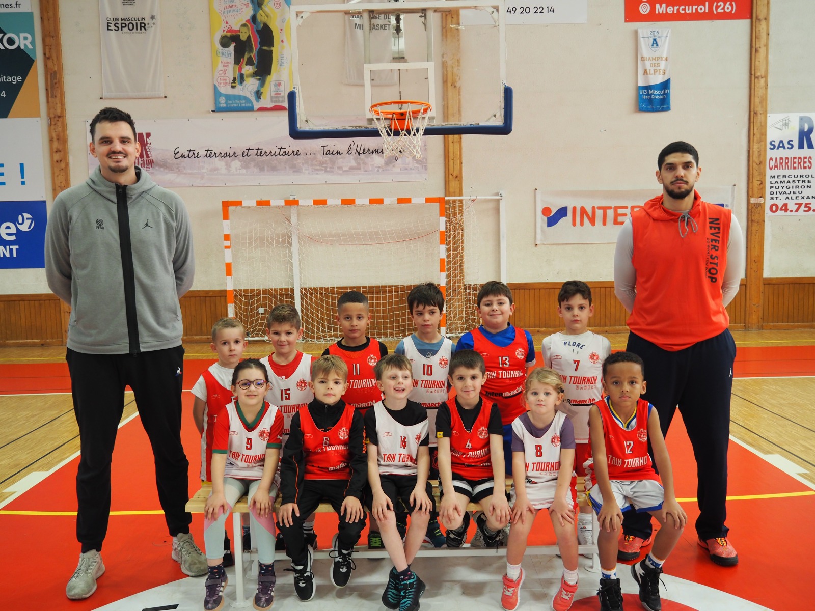 Équipe de basket jeunesse en uniforme posant sur un banc avec deux entraîneurs dans un gymnase.