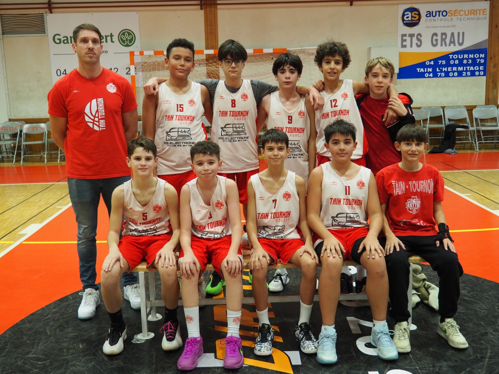 Équipe de jeunes basketteurs posant dans un gymnase avec leur entraîneur en tenues rouges et blanches.
