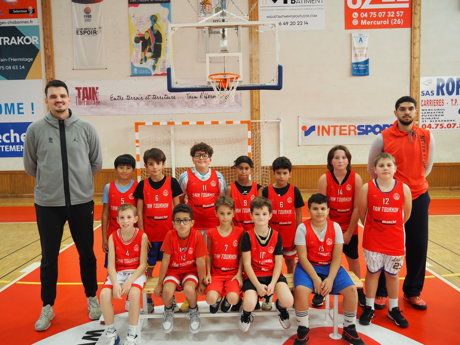 Équipe de basket-ball de jeunes joueurs en maillots rouges posant avec deux entraîneurs en gymnase.