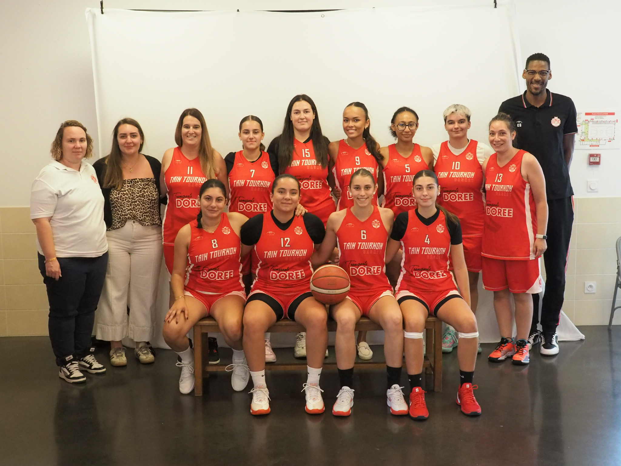 Équipe féminine de basket-ball posant en maillots rouges avec deux entraîneurs debout aux côtés.