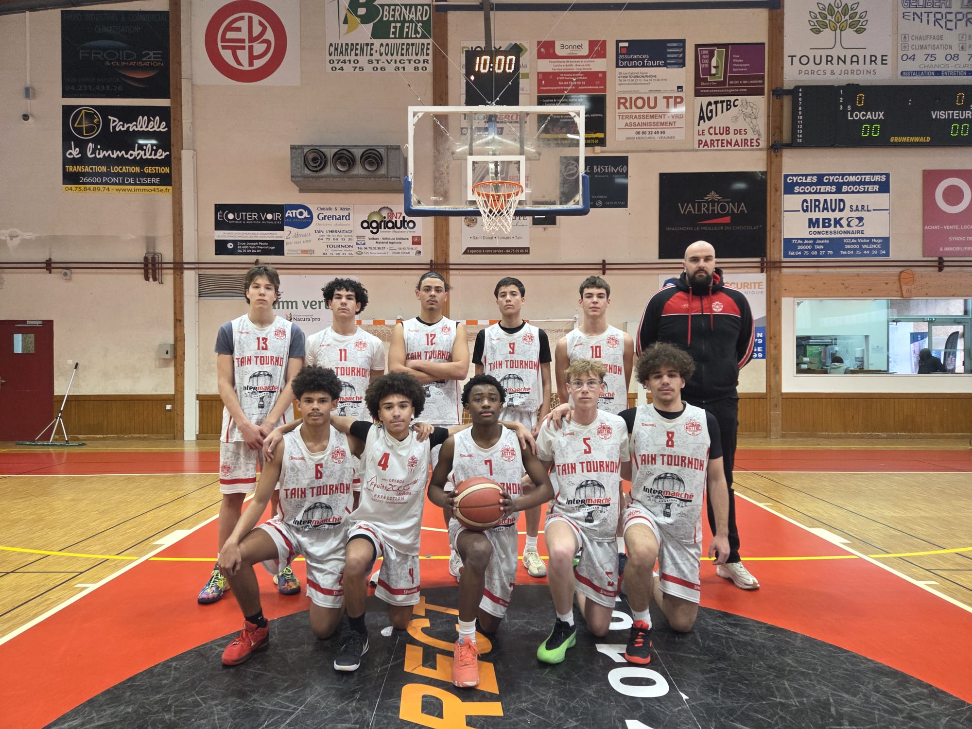 Équipe de basketball juniors posant devant le panier dans un gymnase avec l'entraîneur debout derrière eux.