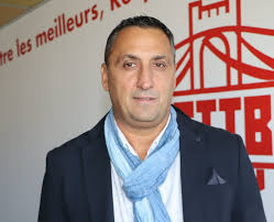 Homme portant une veste noire et une écharpe bleue posant devant un mur avec logo rouge et texte français.
