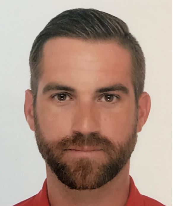 Portrait d'un homme aux cheveux courts, barbe taillée et chemise rouge, regardant l'objectif.