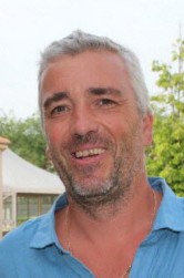 Homme souriant aux cheveux gris et barbe, portant une chemise bleue, avec des arbres en arrière-plan.