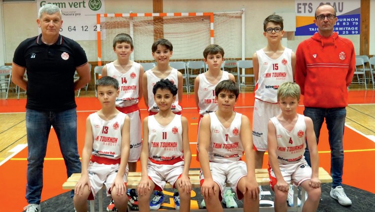 Équipe de jeunes joueurs de basket avec leurs deux entraîneurs dans un gymnase, posant devant un but.