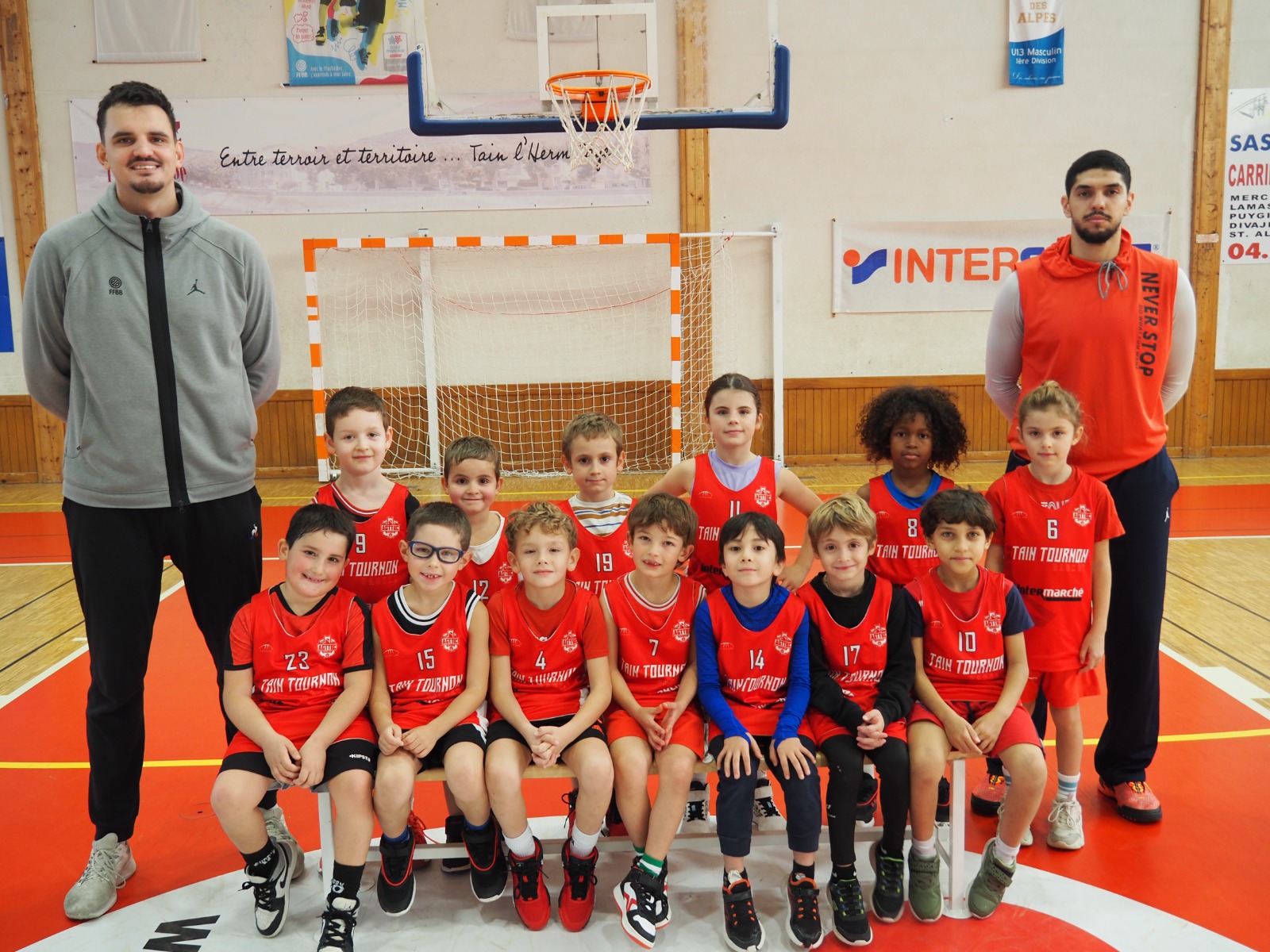 Équipe de jeunes basketteurs et leurs deux entraîneurs posant dans un gymnase devant un panier.