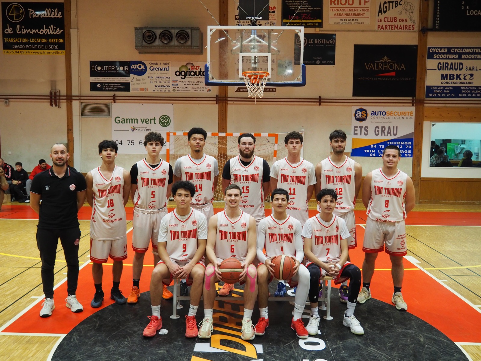 Photo d'équipe de basket en maillots blancs avec coach dans un gymnase, deux joueurs tiennent un ballon.