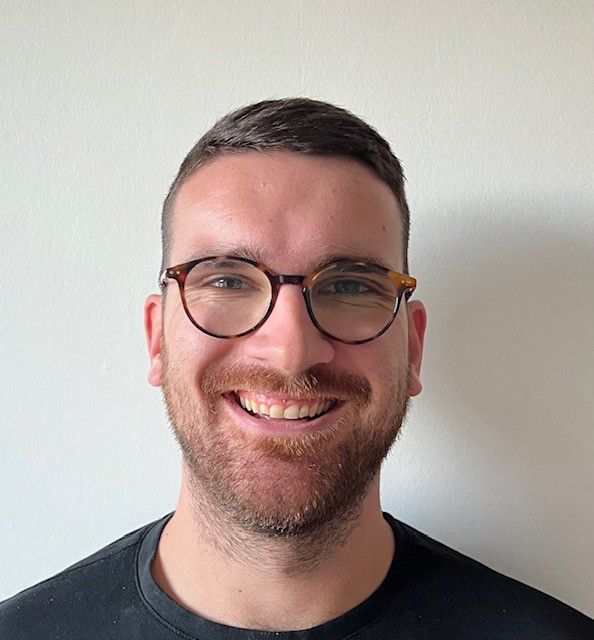 Homme souriant portant des lunettes rondes et une barbe légère devant un mur blanc.