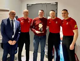 Cinq hommes debout, quatre en uniforme rouge et un en costume sombre, posant pour une photo.