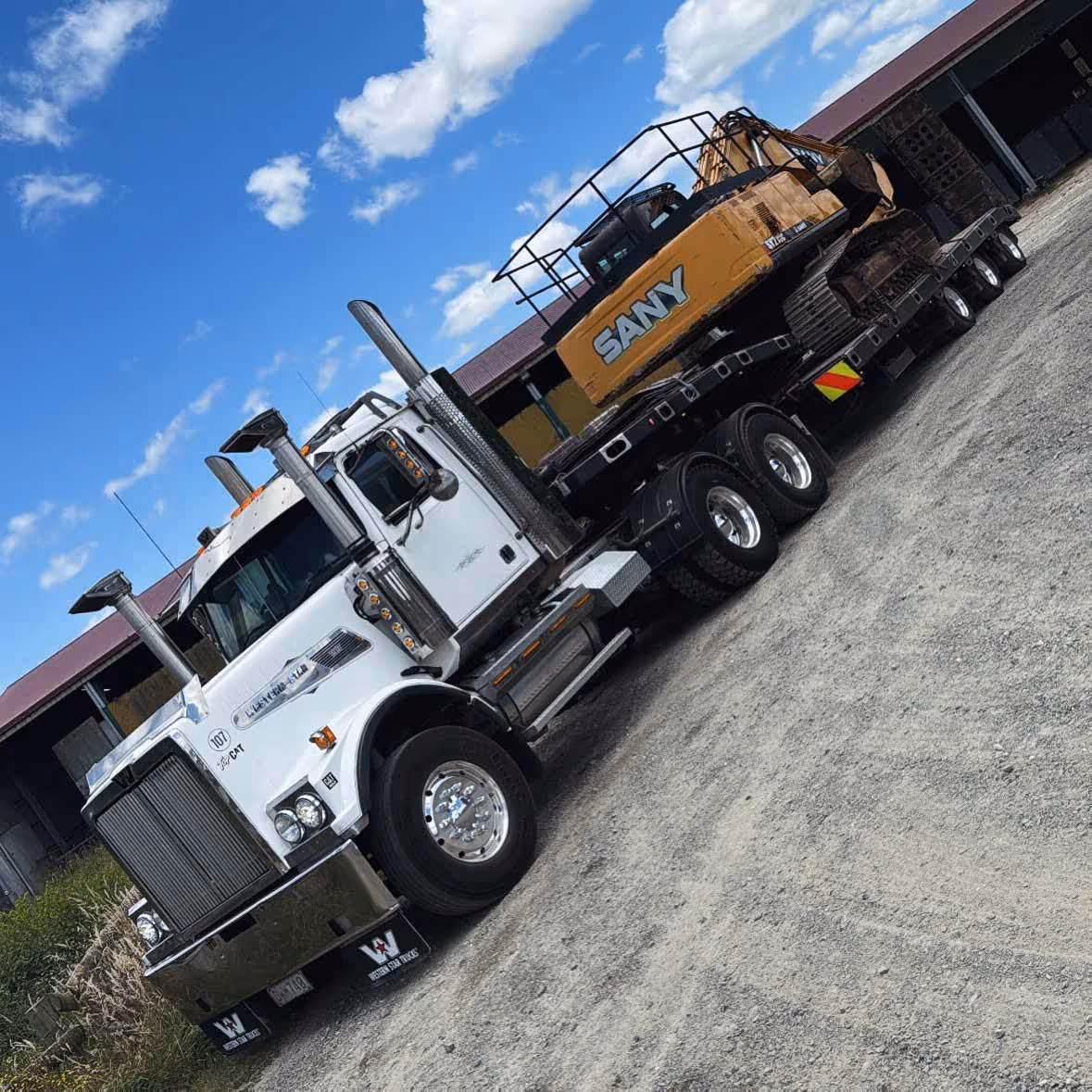 White truck hauling SANY excavator on trailer parked outdoors in a gravel lot.