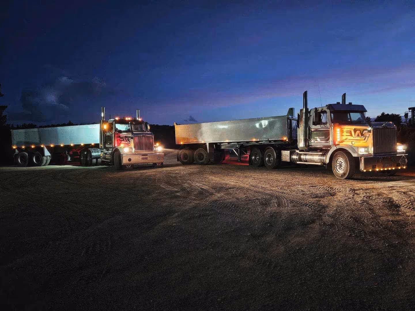 Large semi-trucks with trailers parked on a dirt road at sunset in a rural area.