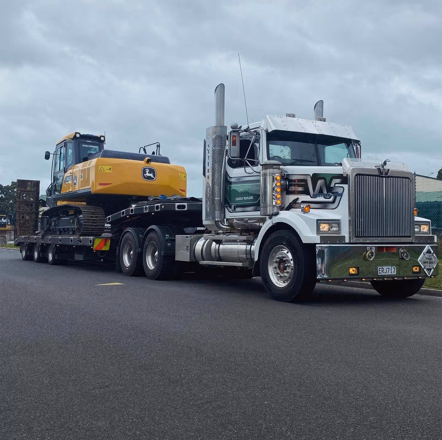 Yellow John Deere excavator transported on trailer by large truck down a city street.