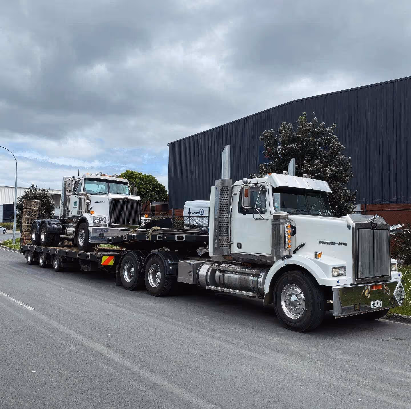 A white truck hauling another vehicle on a flatbed outside a warehouse facility.