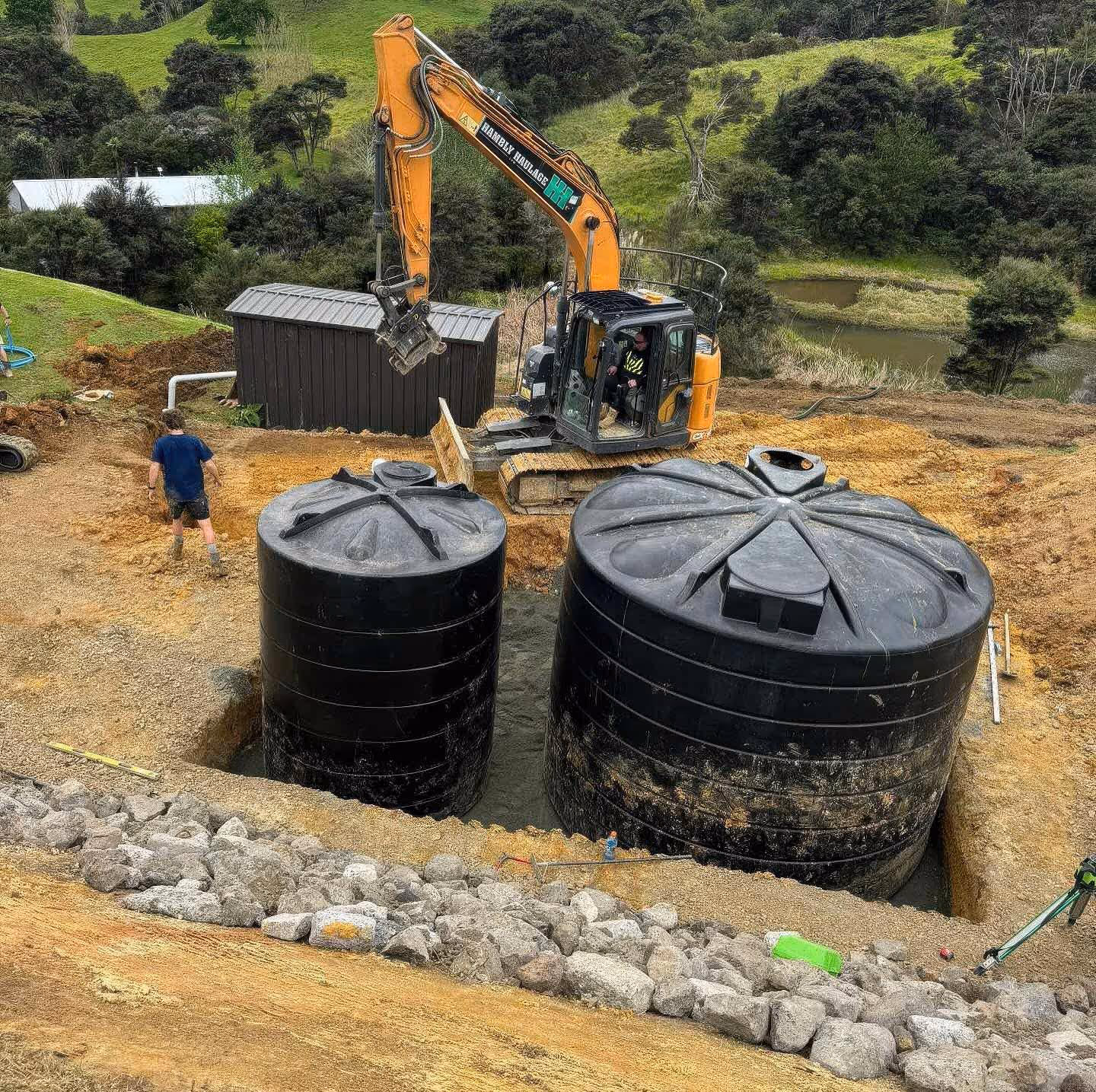 Rural construction site features large black water tanks installed with heavy excavation equipment.