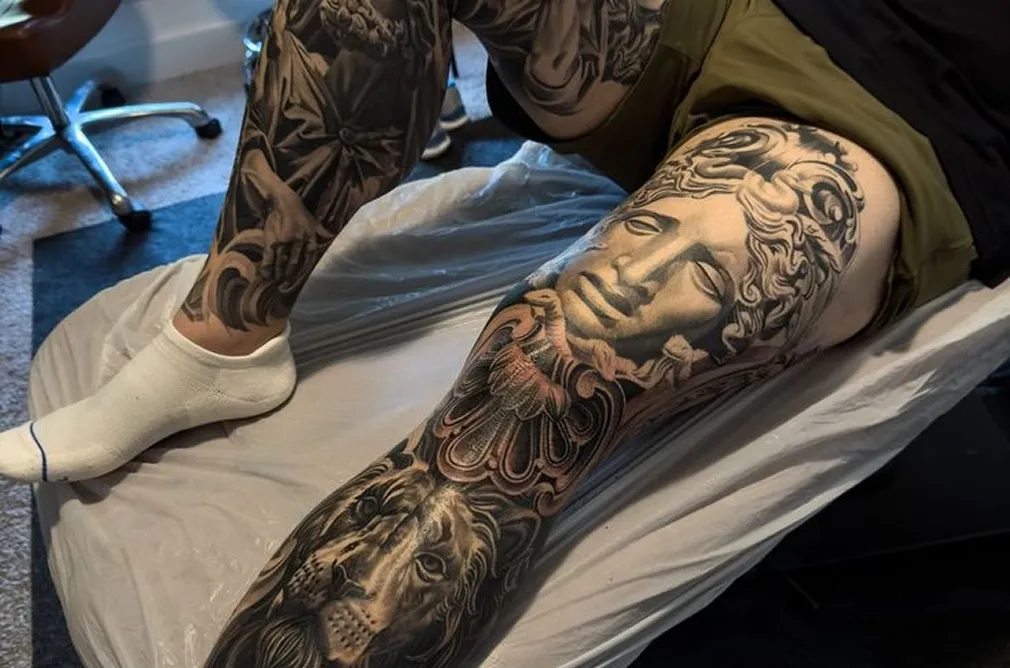 Detailed tattoo on the leg featuring a mix of classical and modern elements, including faces and geometric designs.