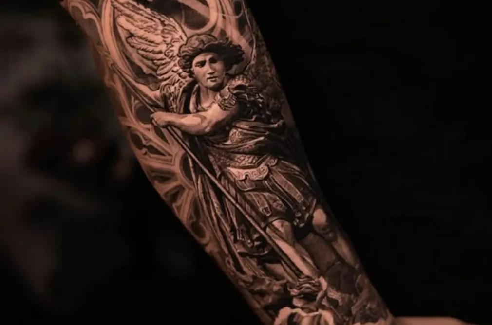 Detailed black and grey tattoo of an angel with wings and a sword, representing a warrior figure on the arm.
