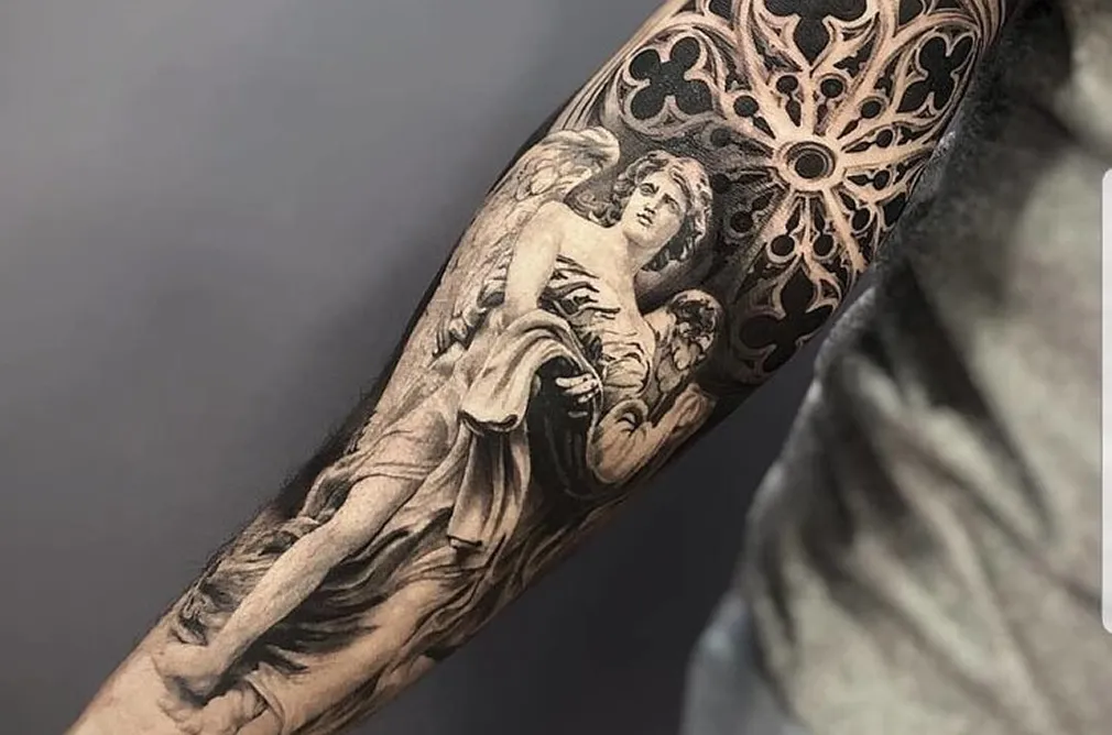 Detailed black and grey tattoo of an angel with wings and intricate geometric patterns on the forearm.
