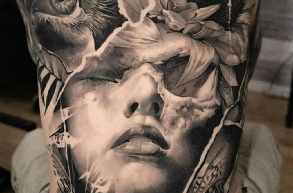Close-up of a black and grey tattoo depicting a shattered face with intricate details of flowers and feathers on the chest