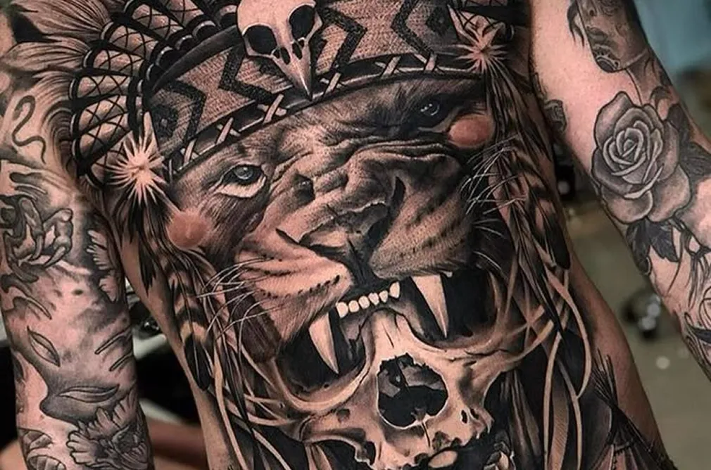 Tattoo of a lion's face and skull, blended into a Native American inspired design on the chest and shoulder.