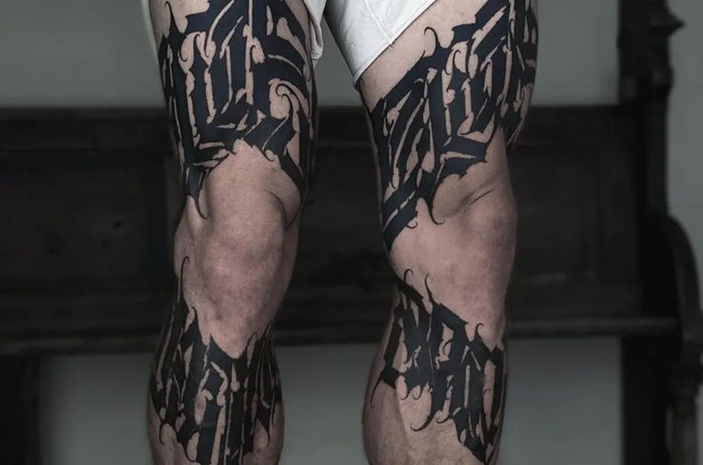 Detailed black and grey tattoo of gothic script and symbols wrapping around the legs, with dark and abstract design.