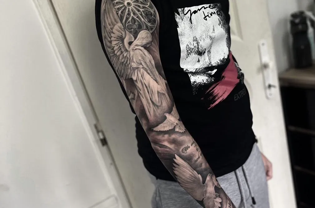Black and grey tattoo featuring an angel with a sword and elaborate background on the arm, showcasing classical art