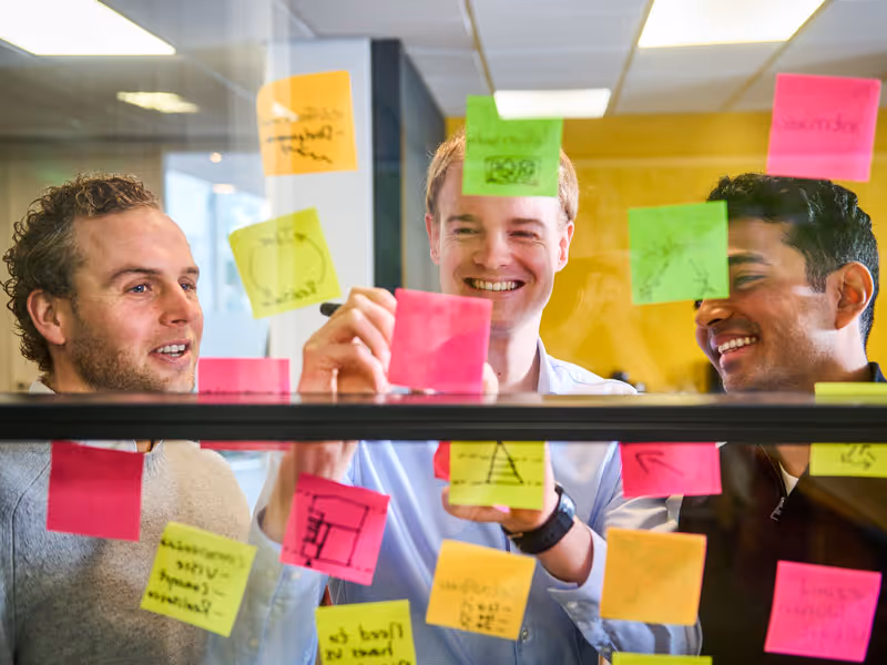 Drie lachende mannen bespreken en schrijven op gekleurde plakbriefjes op een glazen wand in een kantoor.