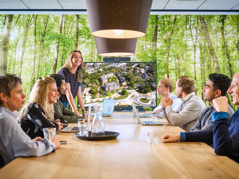 Groep mensen vergadert rond een tafel met laptops, terwijl een vrouw staat en een presentatie geeft met een scherm met luchtfoto van een woonwijk, achter een raam met uitzicht op bomen.
