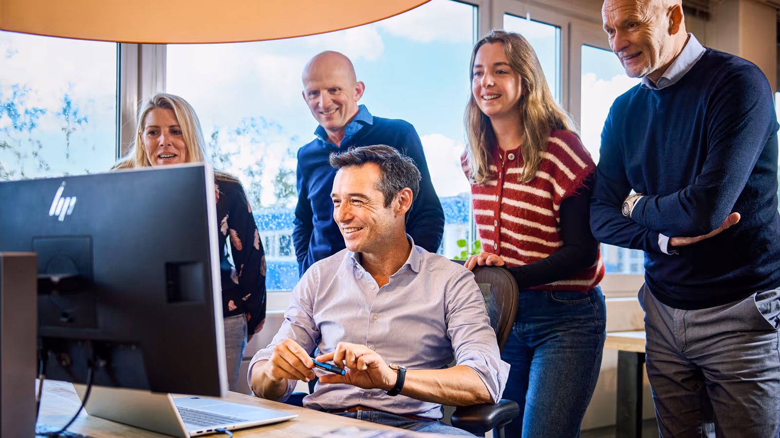 Vijf collega's bekijken samen iets op een computerscherm in een kantooromgeving met ramen op de achtergrond.