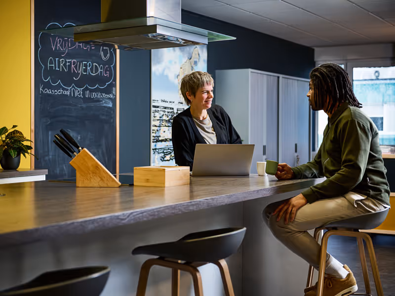 Twee mensen zitten aan een keukenbar, een vrouw kijkt glimlachend naar een man die een kopje vasthoudt.