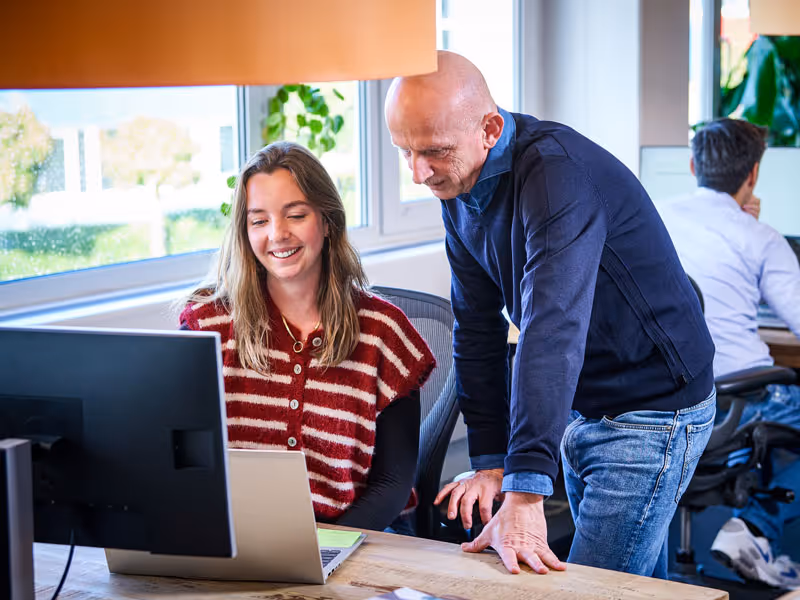 Een man helpt glimlachende vrouw die aan een laptop werkt in een kantoor met grote ramen.