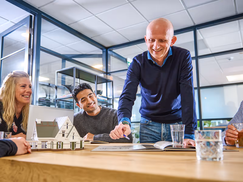 Een groep volwassenen bespreekt bouwplannen rond een tafel met een modelhuis in een modern kantoor.