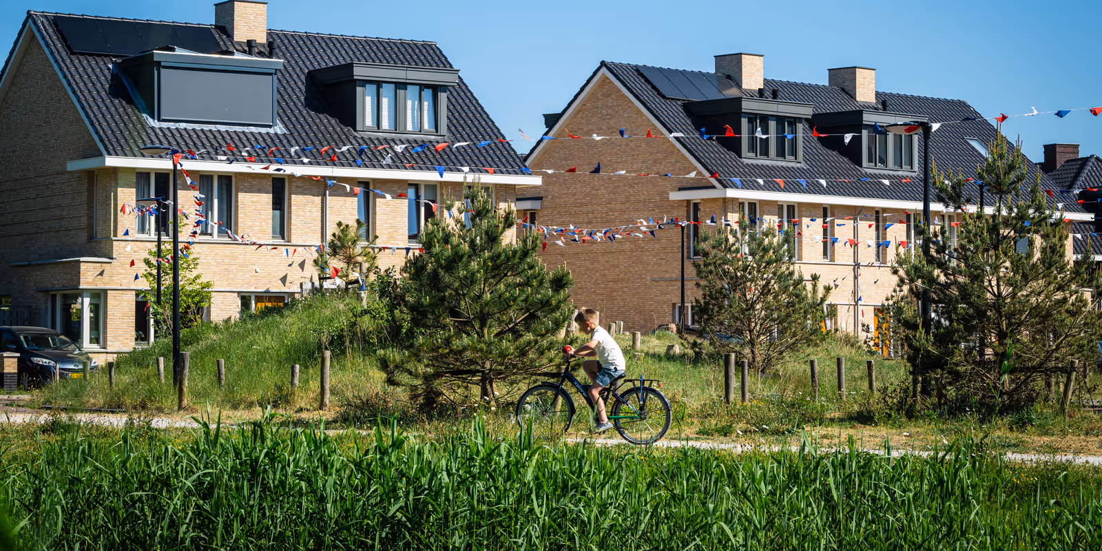Jongen fietst langs groene planten met moderne huizen en kleurrijke vlaggenlijnen op een zonnige dag.