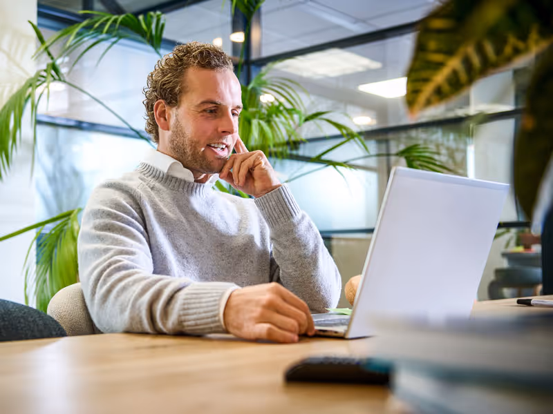 Man met een grijze trui kijkt glimlachend naar een laptop in een kantoorruimte met groene planten op de achtergrond.