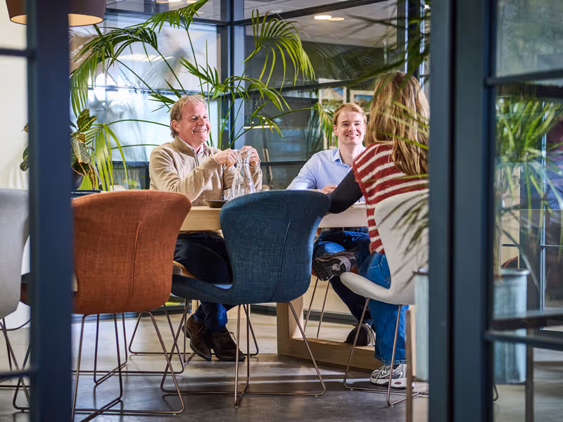 Drie mensen die ontspannen rond een tafel zitten in een moderne, groene kantoorruimte.