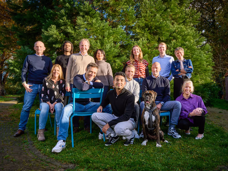 Groep van elf mensen en een hond die buiten op gras voor groene bomen poseren.