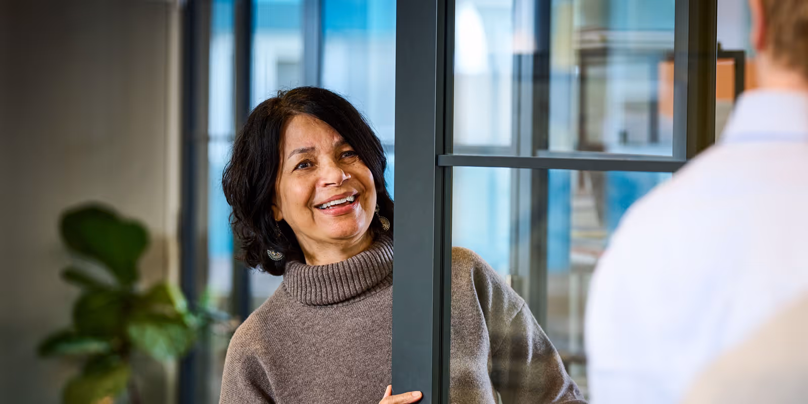 Vrouw in grijze trui glimlacht terwijl ze achter een glazen deur staat en praat met een man in overhemd.