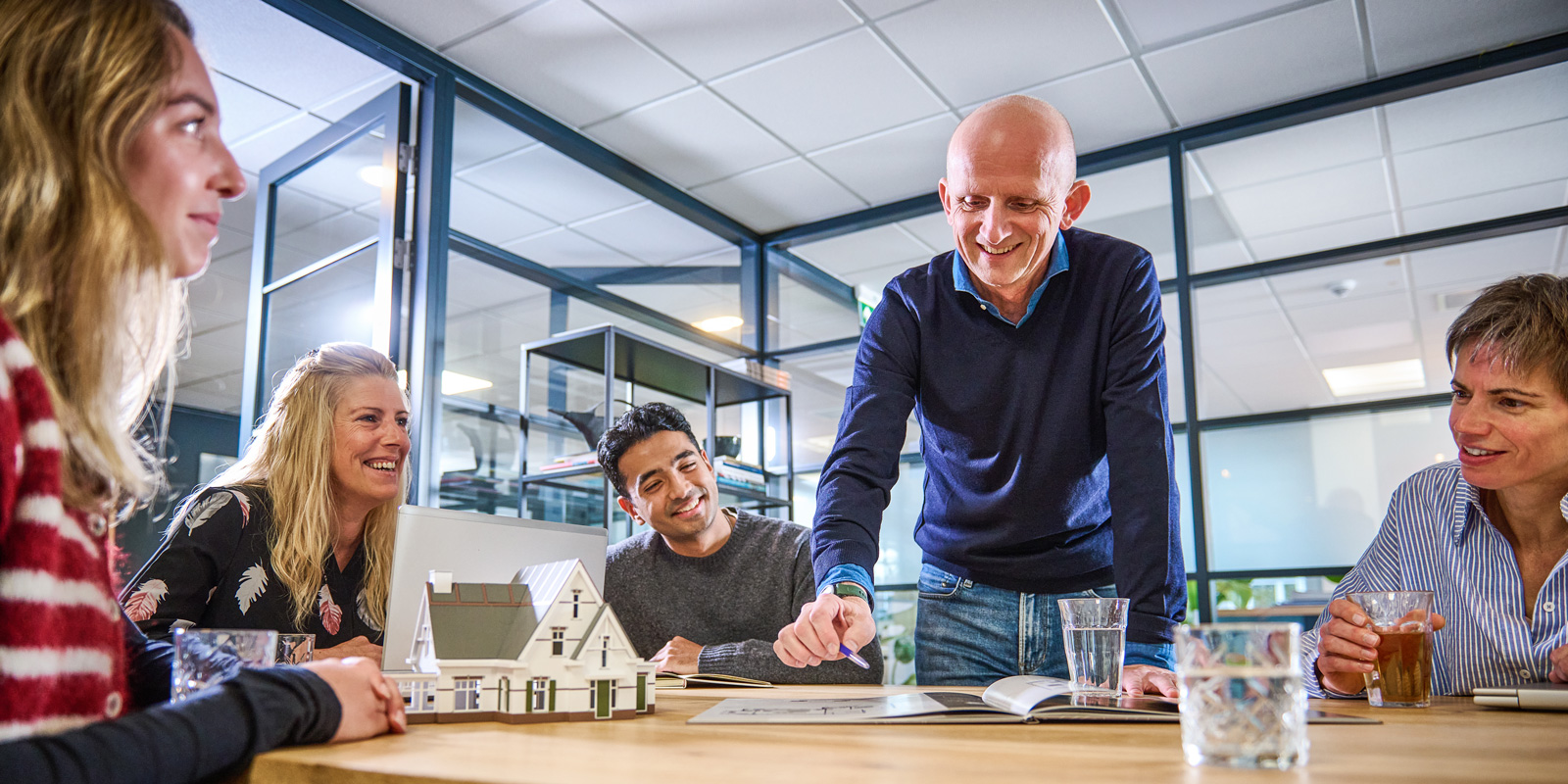 Vijf mensen in een moderne kantoorruimte bespreken een maquette van een huis die op tafel staat.