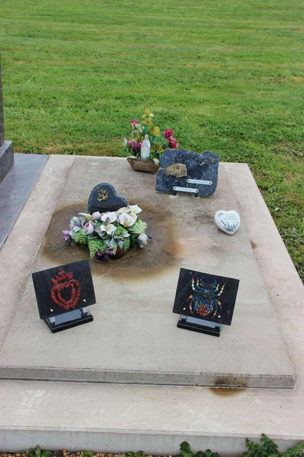 Tombe ornée de plusieurs plaques funéraires, dont un cœur sacré rouge et un scarabée multicolore, avec des fleurs artificielles et une statue religieuse en arrière-plan.