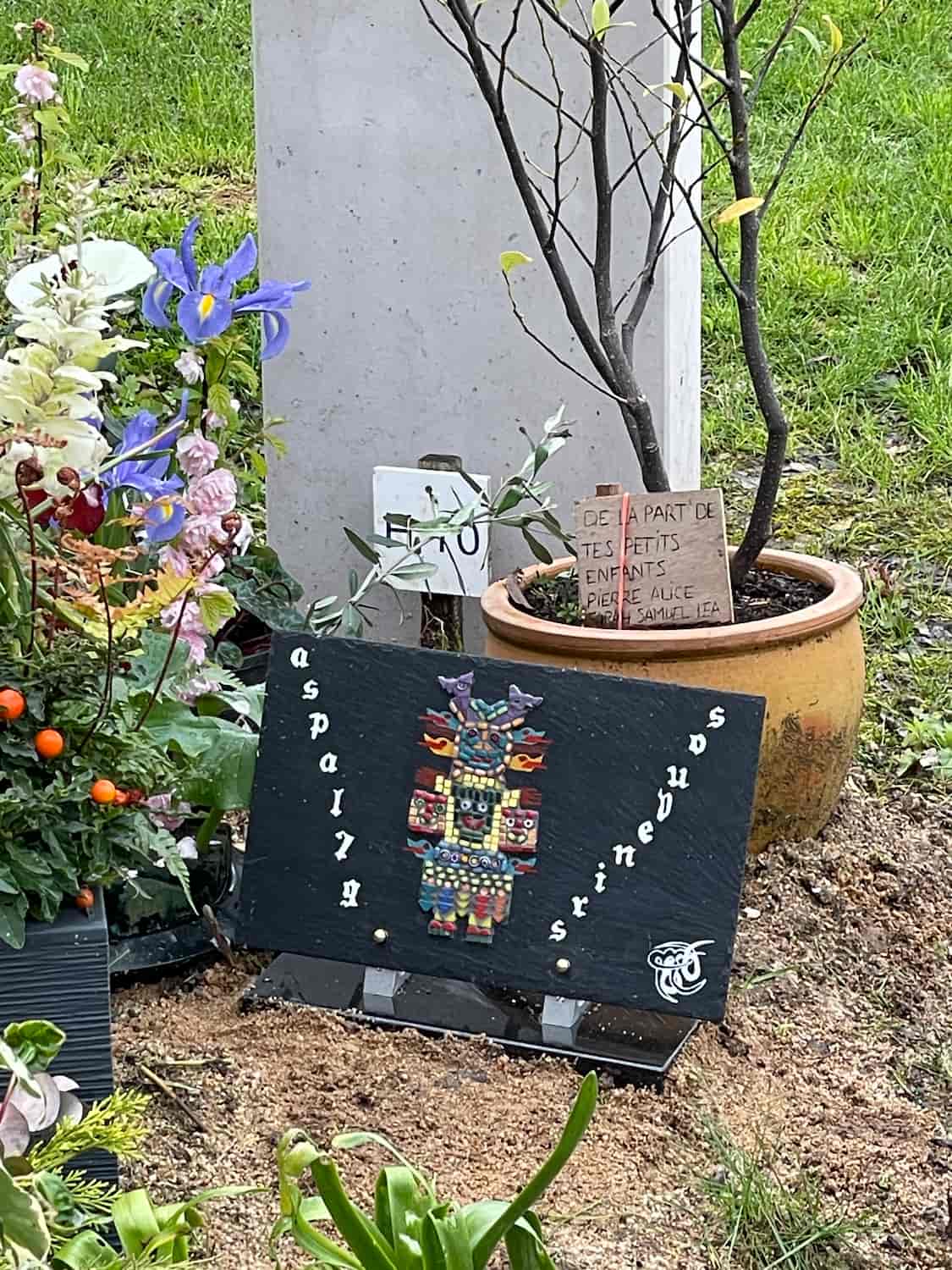 Tombe décorée avec des fleurs, une plaque noire ornée d’un totem coloré et un message manuscrit dans un pot de fleur de la part des petits-enfants.