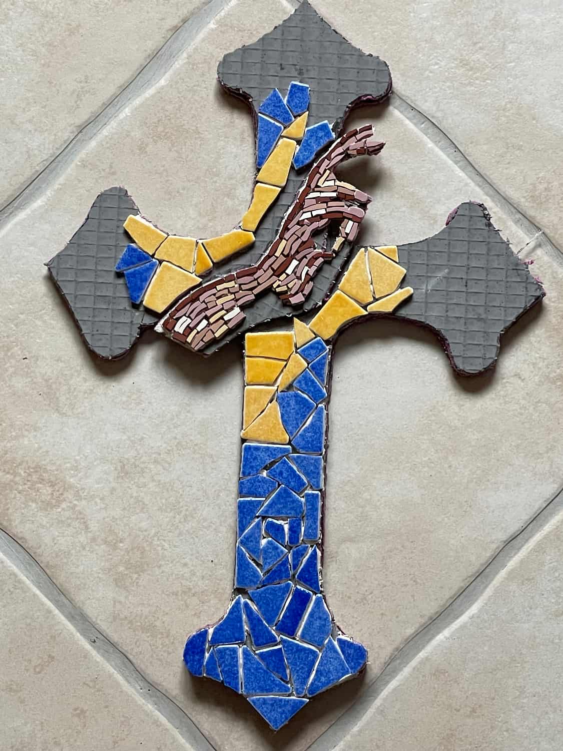 Plaque funéraire en mosaïque représentant une croix aux teintes bleues, orange et marron.