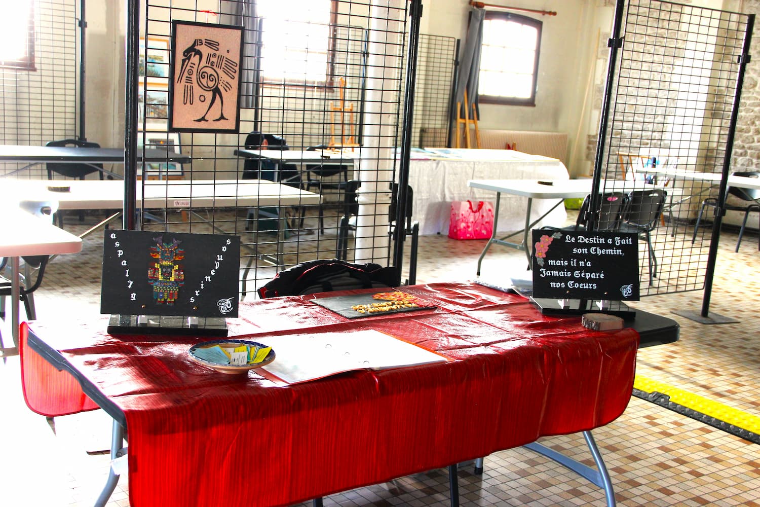 Stand d'exposition artistique avec des tableaux décoratifs, une nappe rouge et des grilles d'affichage dans une salle lumineuse