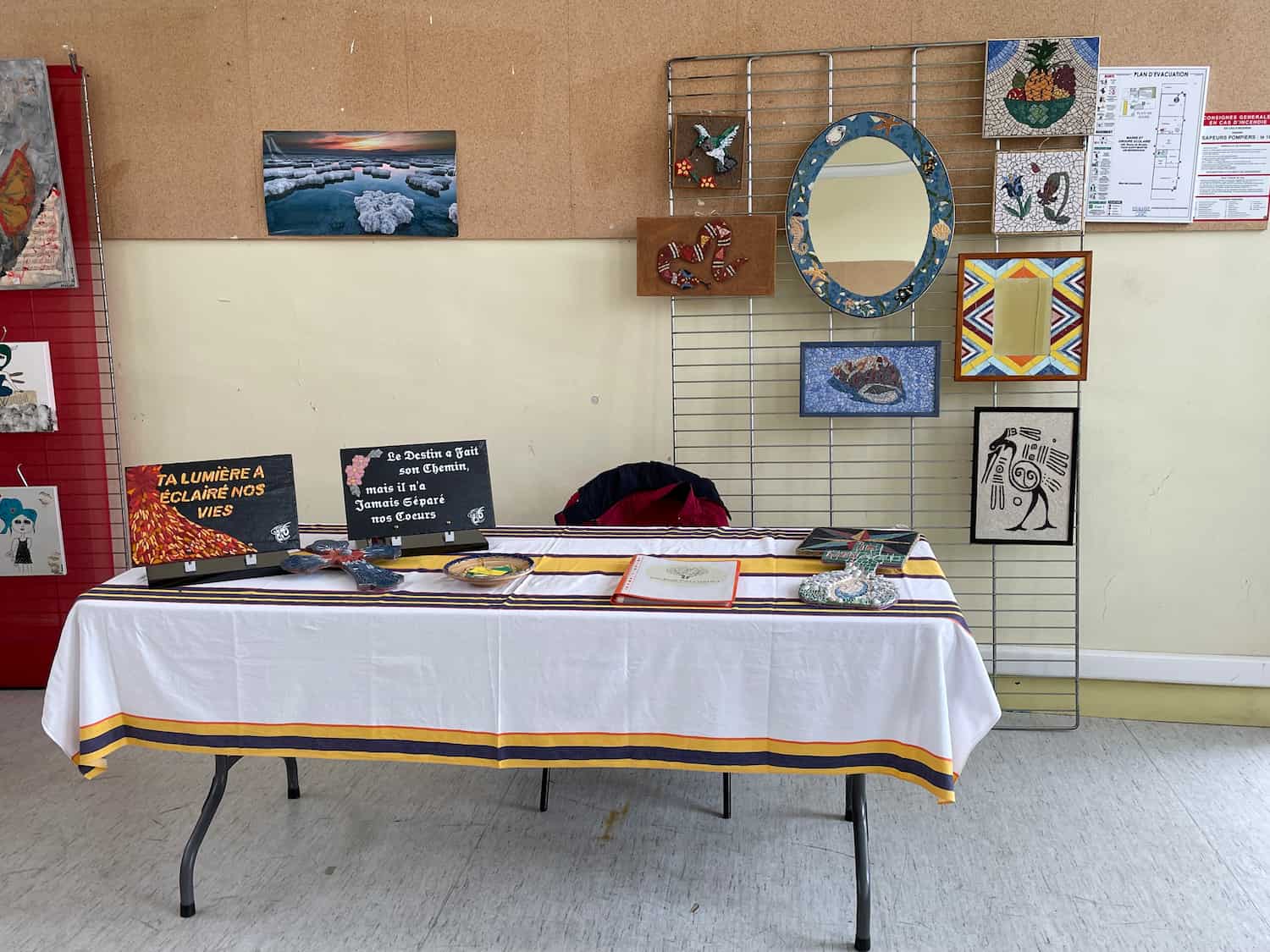 Stand d'exposition présentant des plaques funéraires personnalisées avec des messages émouvants, ainsi que des miroirs et tableaux en mosaïque accrochés sur des grilles.