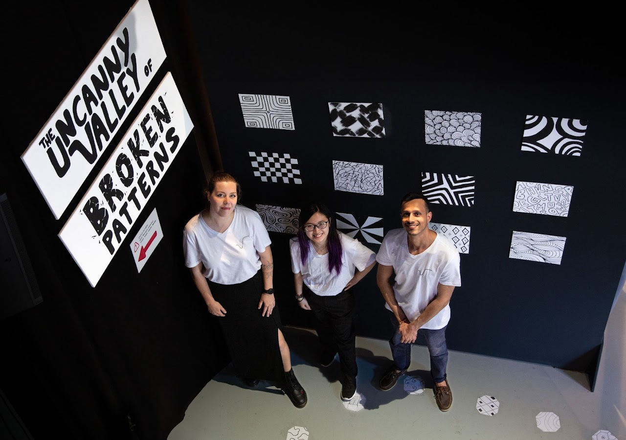 Team picture of three people standing and smiling inside a room with black walls decorated with various black-and-white patterned artworks and a sign reading 'The Uncanny Valley of Broken Patterns.'