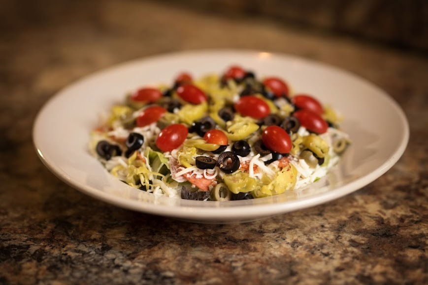 A bowl of salad featuring black olives, diced tomatoes, and crumbled cheese on a wooden table.