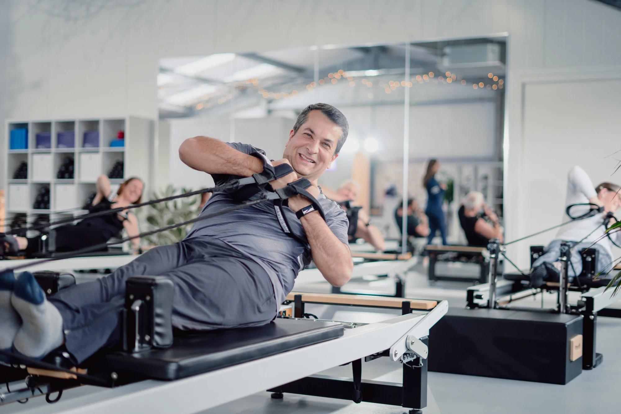 Man doing reformer pilates in a clean studio with mirrors and other people doing reformer pilates.